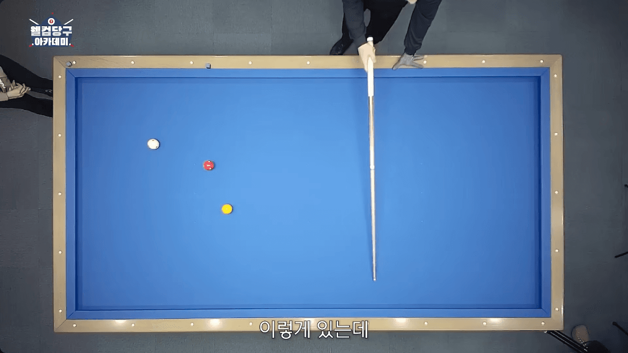 The 7-minute scene from 웰컴 당구아카데미. A white, a red and a yellow ball sit on the left side of the table, and a cue is used to explain the direction.